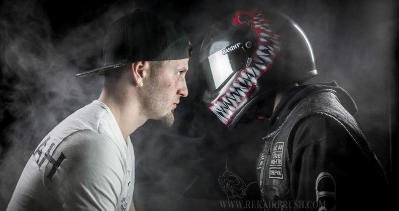 Puede incluir: Un casco de moto negro con un dise&ntilde;o de dientes rojo y blanco amenazante, frente a una persona con una camiseta blanca y una gorra negra. El casco est&aacute; etiquetado como "Bandit". El fondo es oscuro con un efecto de humo.