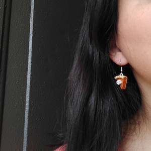 May include: A pair of pie slice earrings with a silver hook. The earrings are brown with a white topping and a silver hook. The earrings are a fun and unique accessory.