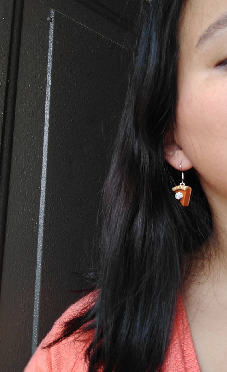 May include: A pair of pie slice earrings with a silver hook. The earrings are brown with a white topping and a silver hook. The earrings are a fun and unique accessory.