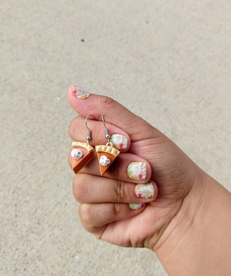 May include: A pair of dangle earrings shaped like pumpkin pie slices. Each earring features a brown crust, orange filling, and a dollop of white whipped cream. The earrings are held by a hand with painted nails.