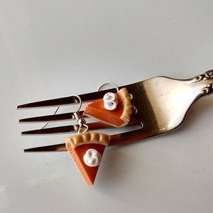 May include: A pair of novelty earrings shaped like pumpkin pie slices. Each earring features an orange pie filling with a tan crust and a dollop of white whipped cream. The earrings are suspended from silver-colored fish hook ear wires.