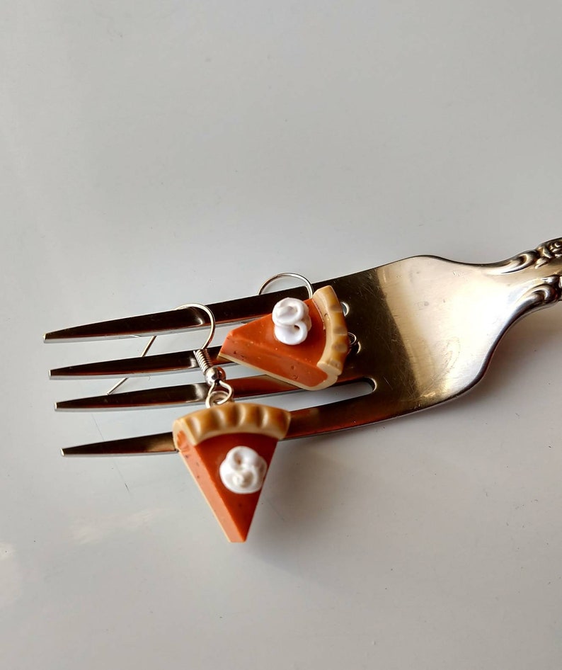 May include: A pair of novelty earrings shaped like pumpkin pie slices. Each earring features an orange pie filling with a tan crust and a dollop of white whipped cream. The earrings are suspended from silver-colored fish hook ear wires.