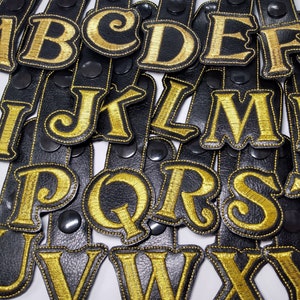 May include: A set of 26 black leather patches with gold embroidered letters, each letter of the alphabet is represented. The patches are stitched with a decorative border.