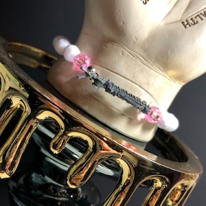 May include: A white and pink beaded bracelet with a silver charm of a spaceship. The bracelet is on a gold-colored stand.