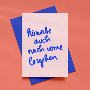 Könnte beinhalten: Eine Grußkarte mit dem deutschen Satz "Könnte auch nach vorne losgehen" in blauer Schrift auf hellrosa Hintergrund. Die Karte liegt auf einem blauen Umschlag, vor korallenfarbenem Hintergrund.