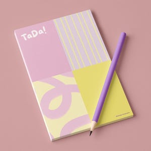 Könnte beinhalten: Ein pastellfarbener Notizblock mit dem Wort "TaDa!" in Weiß und einem lila Bleistift. Der Notizblock hat ein geometrisches Design mit rosa, gelben und hellvioletten Abschnitten. Der Bleistift liegt auf dem Notizblock.