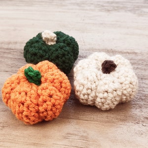 May include: Three crocheted pumpkins in shades of orange, green, and white. The orange pumpkin has a green stem, the green pumpkin has a cream stem, and the white pumpkin has a brown stem. The pumpkins are arranged on a light wood surface.