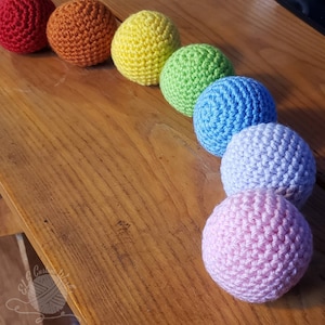 May include: A rainbow of crocheted balls in red, orange, yellow, green, blue, lavender, and pink. The balls are arranged in a semi-circle on a wooden surface.