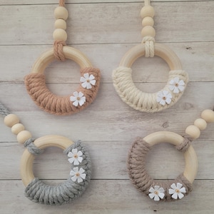 May include: Four wooden rings with macrame detailing and small white flower accents. Each ring is a different colour: light brown, cream, grey, and dark brown. The rings are suspended by a cord with wooden beads.