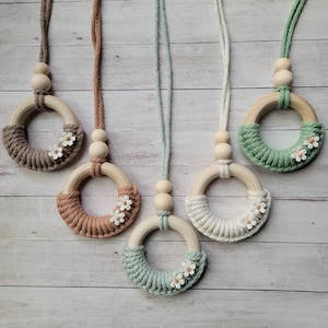 May include: Five wooden ring necklaces with macrame and flower accents. The necklaces are in shades of brown, green, and white.