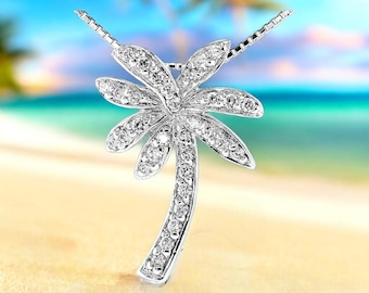 May include: A silver palm tree pendant necklace with a diamond-encrusted design. The necklace is on a white sandy beach with a blue ocean in the background.