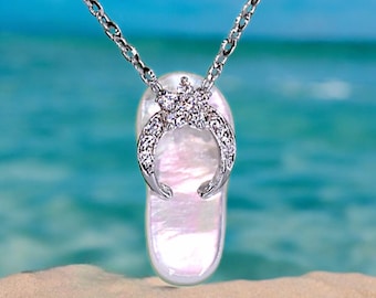 May include: A silver chain necklace with a white seashell pendant. The pendant is shaped like a flip-flop and has a crescent moon design with clear rhinestones.