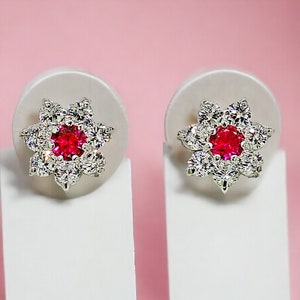 May include: A pair of silver earrings with a red gemstone in the center of each earring. The earrings are shaped like a flower with white gemstones around the red gemstone.