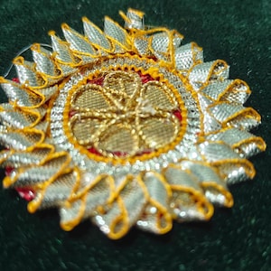 May include: A detailed, circular embellishment with a gold center and silver and gold trim. The design features a floral pattern in the center, surrounded by a red border. The background is a dark green fabric.