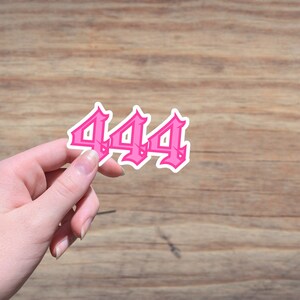 May include: A pink sticker with the number 444 in a gothic font style. The sticker has a white border and is held against a wooden background.
