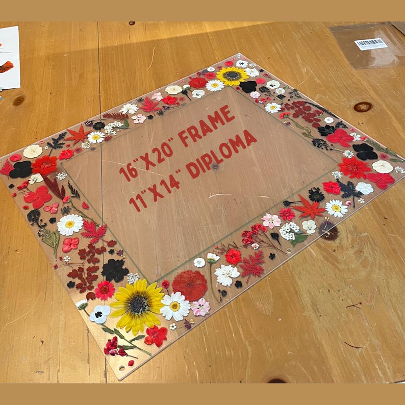 Pode incluir: Uma moldura de acr&iacute;lico transparente com uma borda floral. A moldura &eacute; projetada para uma moldura de 40,6 cm x 50,8 cm e um diploma de 27,9 cm x 35,6 cm. A borda apresenta flores secas coloridas, incluindo girass&oacute;is e margaridas.