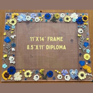 Pode incluir: Uma moldura retangular com fundo de madeira, adornada com uma variedade de flores secas coloridas. A moldura foi projetada para conter uma foto ou obra de arte de 28 cm x 36 cm, com espa&ccedil;o para um diploma de 22 cm x 28 cm. As flores incluem flores amarelas, azuis e brancas.