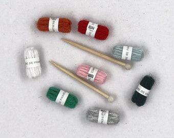 May include: A collection of miniature knitting supplies, including small yarn skeins in various colors like red, orange, and green, along with two wooden knitting needles. Each yarn skein has a white label.