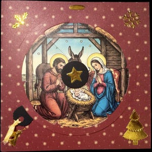 May include: A square maroon card featuring a circular cutout displaying a nativity scene with Joseph, Mary, and the infant Jesus in a manger. The card is decorated with gold foil accents including a star, a snowflake, a tree, and holly leaves.