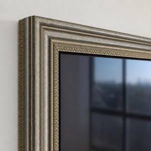 Op de afbeelding: Een rechthoekige spiegel met een brede, sierlijke lijst met een metallic zilveren en bronzen afwerking. De lijst heeft een gedetailleerd, herhalend reliëfpatroon langs de binnenrand, rondom een glad, reflecterend glazen oppervlak.