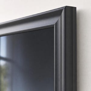 Può includere: Una cornice portafoto nero opaco con un profilo arrotondato liscio e un giunto angolare a 45 gradi. La cornice presenta un bordo interno profondo e incassato che racchiude una superficie scura e riflettente, creando un bordo semplice.
