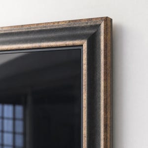 Op de afbeelding: Een rechthoekige wandspiegel met een brede, sierlijke lijst in een verweerde brons- en antracietzwarte afwerking. De lijst heeft een getrapt, geprofileerd ontwerp met een gladde binnenrand en een textuurrijke, metalen buitenrand.