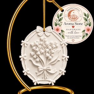 May include: An oval white aroma stone featuring a relief of lily of the valley flowers, suspended by a gold cord from a gold metal stand. A round tag attached to the cord reads: Beautiful Creations, Aroma Stone, HANDMADE with love.