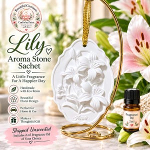 May include: A white oval aroma stone sachet featuring a raised lily flower relief, suspended by a gold ribbon on a gold metal stand. Beside it sits a small brown glass bottle labeled Fragrance Oil 2 ml. The text reads Lily Aroma Stone Sachet.