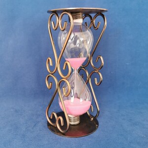 Op de afbeelding: Een zandloper met een heldere glazen bol gevuld met roze zand, geplaatst in een decoratief bronskleurig metalen frame. Het frame heeft sierlijke, wervelende ontwerpen en een cirkelvormige basis. De zandloper is een decoratief item.