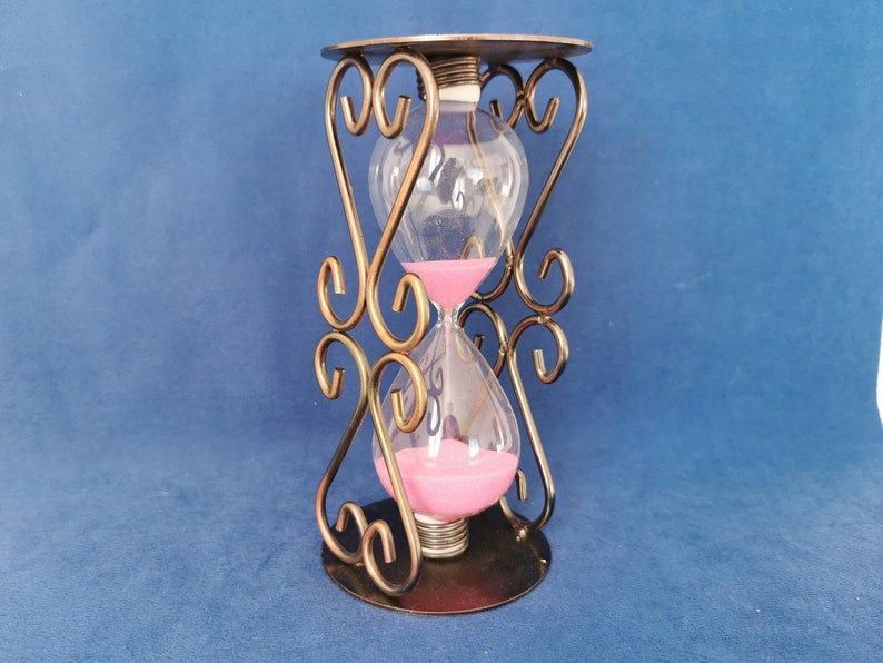 Op de afbeelding: Een zandloper met een heldere glazen bol gevuld met roze zand, geplaatst in een decoratief bronskleurig metalen frame. Het frame heeft sierlijke, wervelende ontwerpen en een cirkelvormige basis. De zandloper is een decoratief item.
