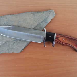 May include: A fixed-blade knife with a silver-toned steel blade featuring the text SA63, a double-sided metal guard, and a handle constructed from a black bolster and a reddish-brown wood grain grip secured with three small metal rivets.