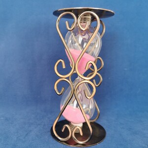 Op de afbeelding: Een zandloper met een heldere glazen bol gevuld met roze zand, geplaatst in een decoratief metalen frame. Het frame heeft sierlijke, wervelende ontwerpen en een cirkelvormige basis en bovenkant. De achtergrond is effen blauw.
