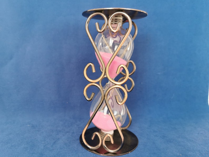 Op de afbeelding: Een zandloper met een heldere glazen bol gevuld met roze zand, geplaatst in een decoratief metalen frame. Het frame heeft sierlijke, wervelende ontwerpen en een cirkelvormige basis en bovenkant. De achtergrond is effen blauw.