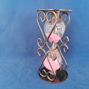 Op de afbeelding: Zandloper met roze zand, ingesloten in helder glas. De zandloper wordt ondersteund door een decoratief metalen frame met hartvormige accenten. De basis en de bovenkant zijn cirkelvormig en het frame heeft een metallic afwerking.