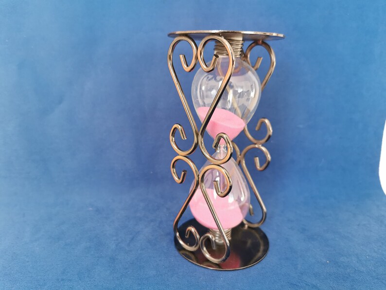 Op de afbeelding: Zandloper met roze zand, ingesloten in helder glas. De zandloper wordt ondersteund door een decoratief metalen frame met hartvormige accenten. De basis en de bovenkant zijn cirkelvormig en het frame heeft een metallic afwerking.