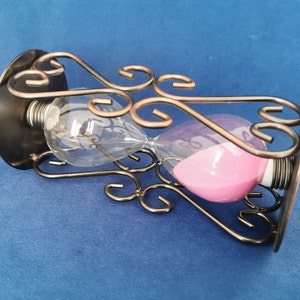 Op de afbeelding: Zandloper met roze zand in een heldere glazen bol, ondersteund door een decoratief bronzen metalen frame. Het frame heeft sierlijke, wervelende ontwerpen. De zandloper staat tegen een blauwe achtergrond.