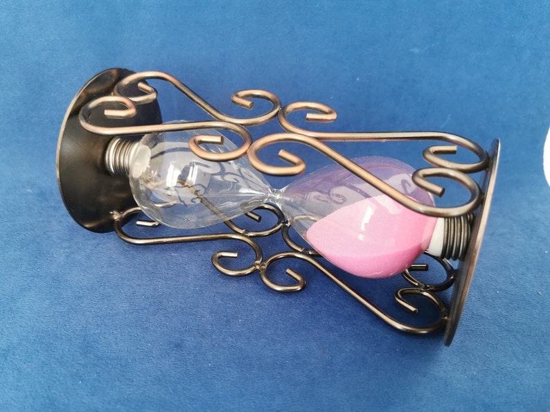 Op de afbeelding: Zandloper met roze zand in een heldere glazen bol, ondersteund door een decoratief bronzen metalen frame. Het frame heeft sierlijke, wervelende ontwerpen. De zandloper staat tegen een blauwe achtergrond.