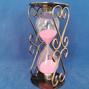Op de afbeelding: Een sierlijke zandloper met roze zand in heldere glazen bollen, ondersteund door een decoratief metalen frame. Het frame heeft een cirkelvormige basis en bovenkant, met hartvormige ontwerpen. Een decoratief item.