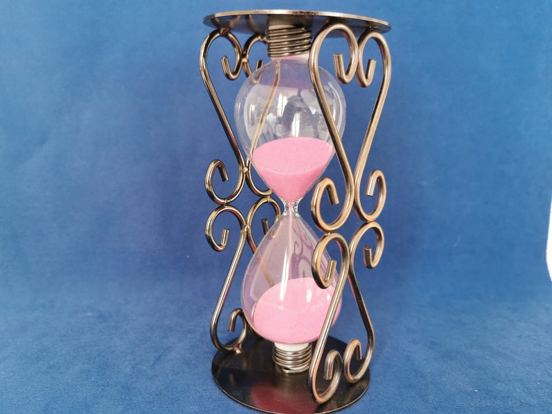 Op de afbeelding: Een sierlijke zandloper met roze zand in heldere glazen bollen, ondersteund door een decoratief metalen frame. Het frame heeft een cirkelvormige basis en bovenkant, met hartvormige ontwerpen. Een decoratief item.