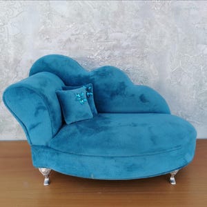 Puede incluir: Una caja de joyería de terciopelo turquesa con forma de chaise longue y una almohada a juego. La caja de joyería tiene patas plateadas y está colocada sobre un fondo gris claro texturizado. La almohada está decorada con adornos azules.