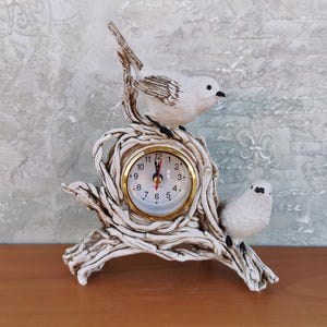 Op de afbeelding: Witte keramische klok met een vogelontwerp. De klok heeft een wit gezicht met zwarte cijfers en wijzers. De klok staat op een houten oppervlak.
