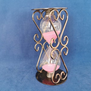 Op de afbeelding: Een zandloper met een heldere glazen behuizing en roze zand. De zandloper wordt op zijn plaats gehouden door een decoratief goudkleurig metalen frame met hartvormige ontwerpen. De basis en de bovenkant zijn cirkelvormig.