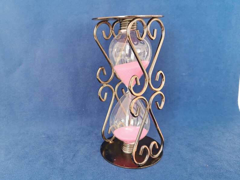 Op de afbeelding: Een zandloper met een heldere glazen behuizing en roze zand. De zandloper wordt op zijn plaats gehouden door een decoratief goudkleurig metalen frame met hartvormige ontwerpen. De basis en de bovenkant zijn cirkelvormig.