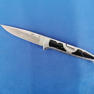 May include: A stainless steel folding knife with a black and silver handle. The blade is engraved with the word "Columbia". The knife is on a blue surface.
