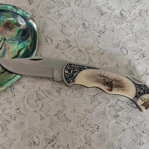 Puede incluir: Una navaja plegable con hoja de metal plateado y mango con incrustación color hueso que presenta un gráfico detallado de una cabeza de ciervo. El mango incluye grabados negros intrincados en ambos extremos y un pequeño detalle de ciervo.