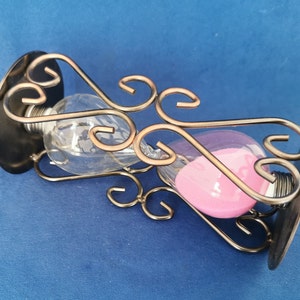 Op de afbeelding: Een zandloper met heldere glazen bollen en roze zand, gehouden in een decoratief metalen frame. Het frame heeft wervelende patronen en wordt ondersteund door twee zwarte cirkelvormige bases. Ongeveer 15 cm hoog.