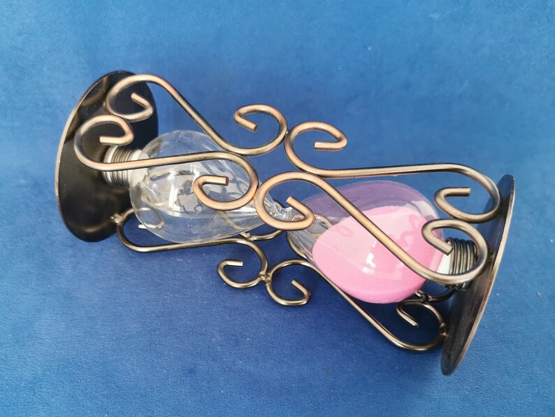 Op de afbeelding: Een zandloper met heldere glazen bollen en roze zand, gehouden in een decoratief metalen frame. Het frame heeft wervelende patronen en wordt ondersteund door twee zwarte cirkelvormige bases. Ongeveer 15 cm hoog.