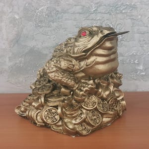 Può includere: Una statua di rana dorata con occhi rossi e una moneta in bocca. La rana è seduta su un mucchio di monete d'oro. La statua ha una superficie strutturata. Lo sfondo è un muro strutturato.