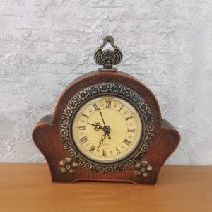 Puede incluir: Un reloj de madera marrón con una esfera dorada y números romanos. El reloj tiene un diseño decorativo alrededor de la esfera y una pequeña manija adornada en la parte superior.