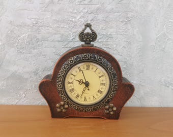 Puede incluir: Un reloj de madera marrón con una esfera dorada y números romanos. El reloj tiene un diseño decorativo alrededor de la esfera y una pequeña manija adornada en la parte superior.